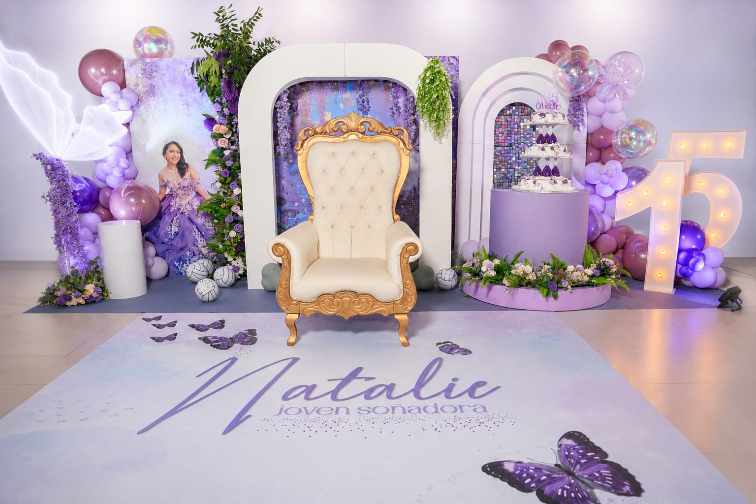 Set completo de XV años con temática de mariposas en tonos lila, trono dorado, torre de pastel y letra luminosa 15